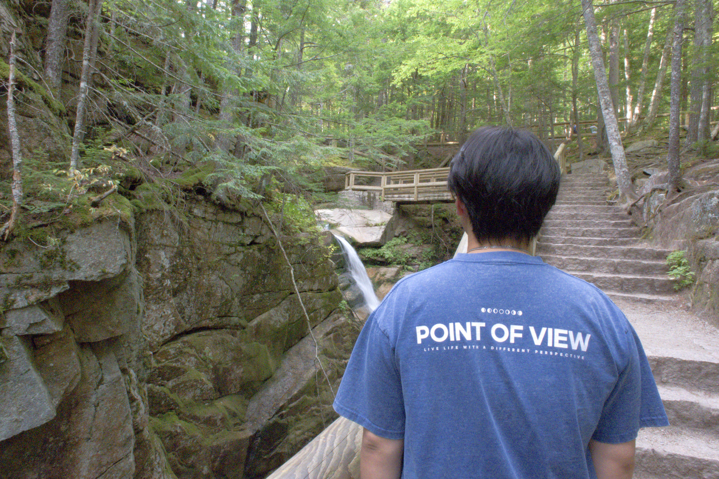 Person wearing a blue t-shirt with 'POINT OF VIEW LIVE LIFE WITH A DIFFERENT PERSPECTIVE' on the back. Standing in nature.