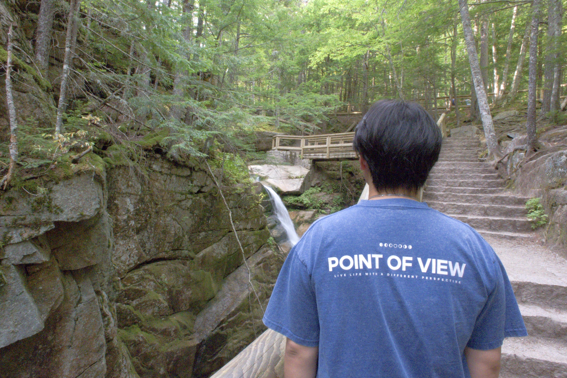 Person wearing a blue t-shirt with 'POINT OF VIEW LIVE LIFE WITH A DIFFERENT PERSPECTIVE' on the back. Standing in nature.