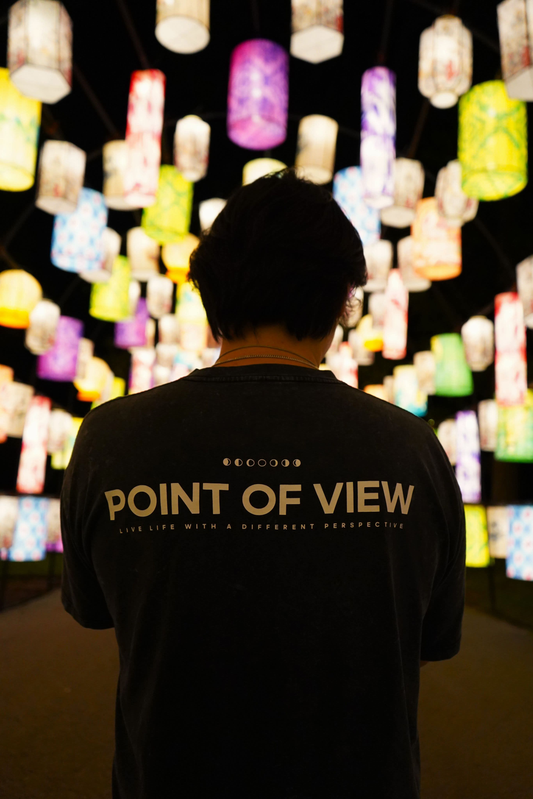 Person wearing a black t-shirt with 'POINT OF VIEW LIVE LIFE WITH A DIFFERENT PERSPECTIVE' text, standing in front of colorful light display.